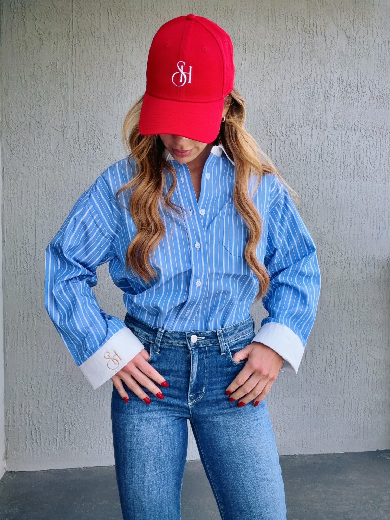 Person wearing a red cap with a logo, blue striped shirt, and jeans against a gray wall.