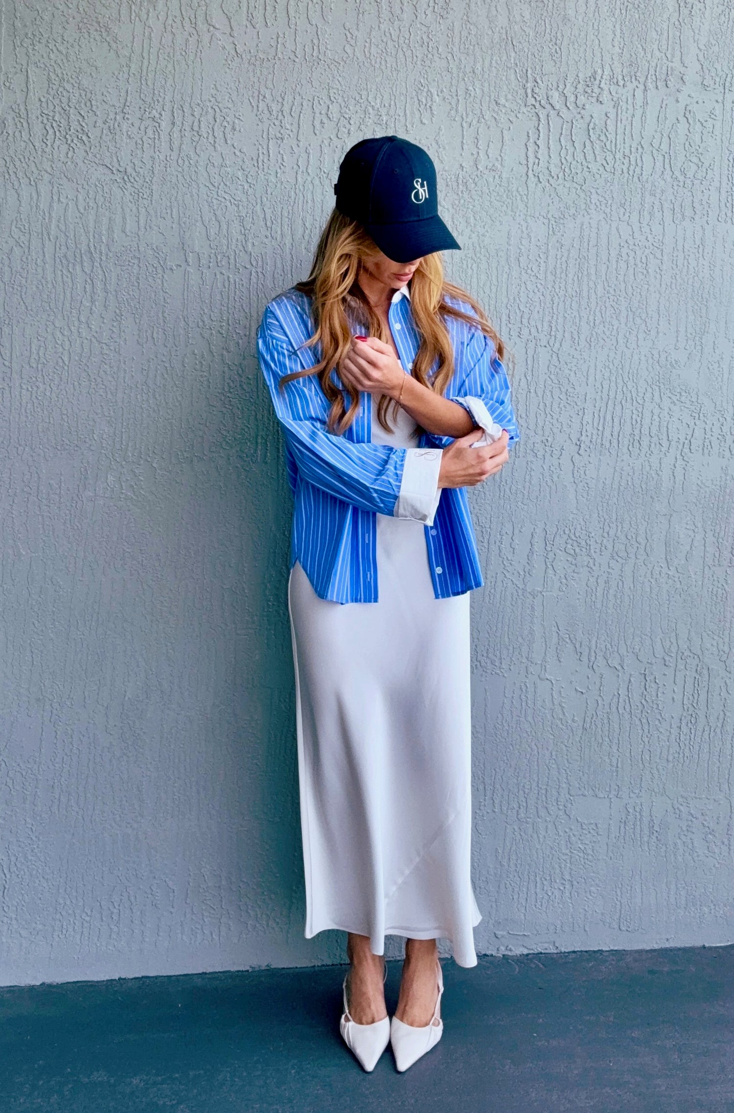Woman wearing a blue shirt, white skirt, and black cap against a gray wall.