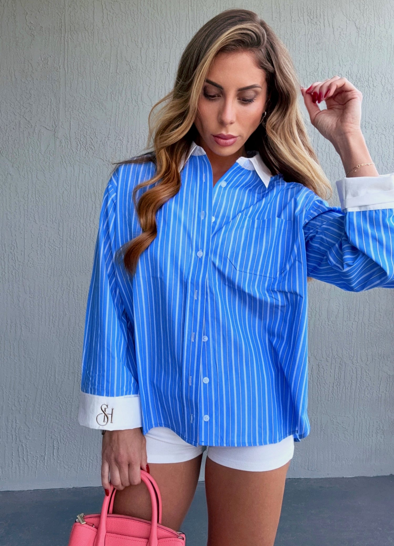 Woman wearing a blue striped shirt with white trim, holding a pink handbag against a gray wall.