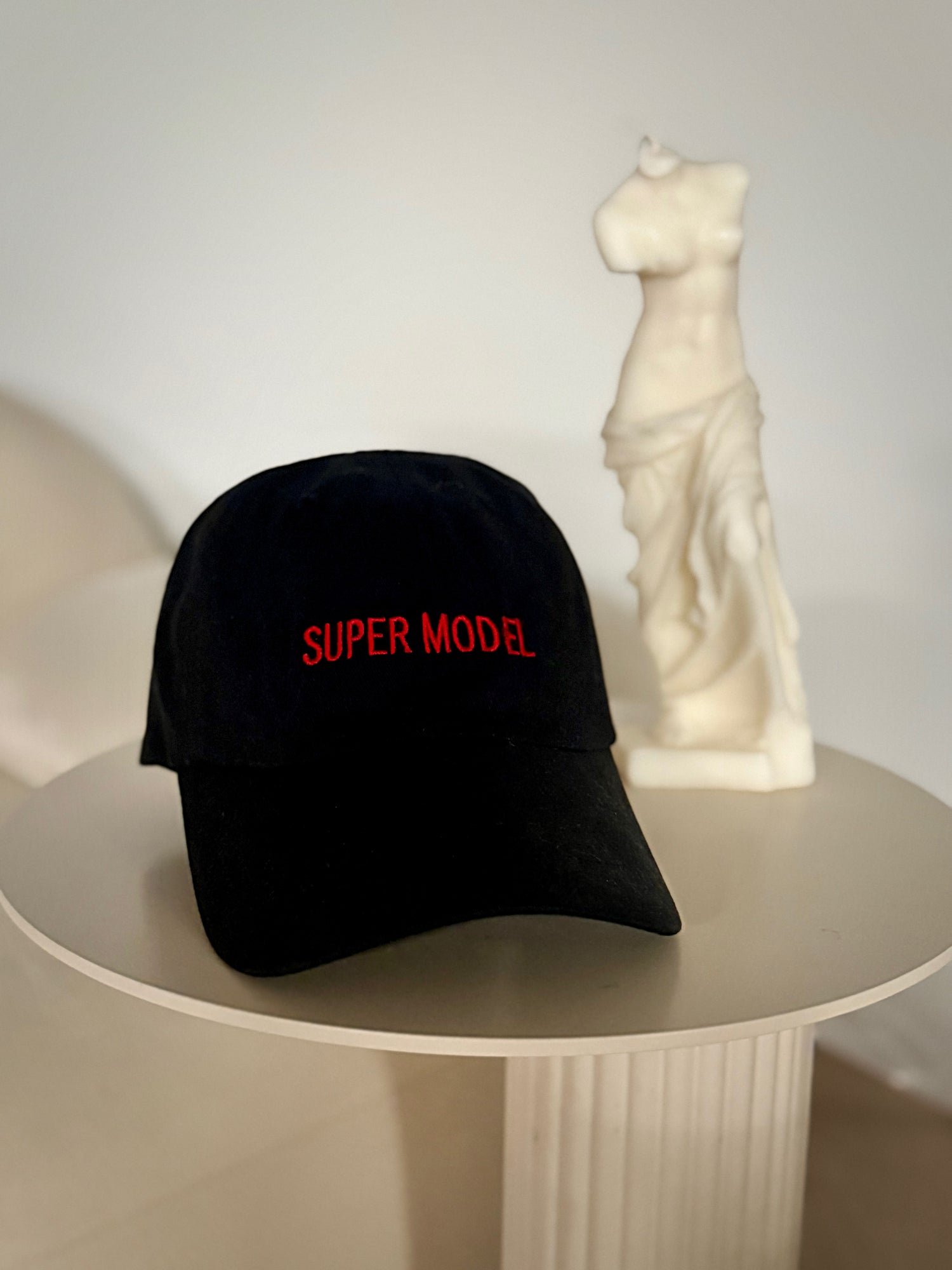 Black cap with 'SUPER MODEL' in red on a white surface with a statue in the background