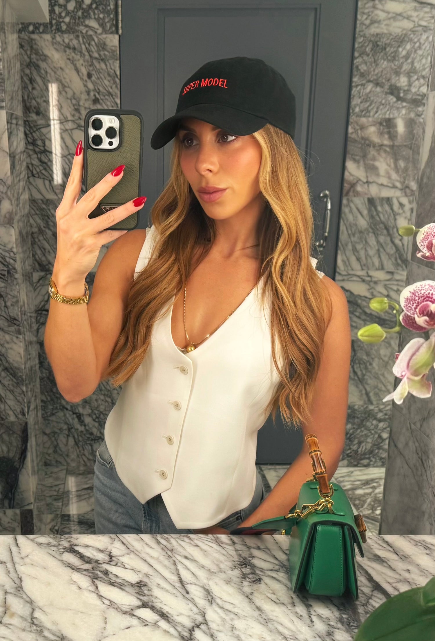 Woman taking a mirror selfie wearing a white blouse and black cap, with a green handbag on a marble surface.