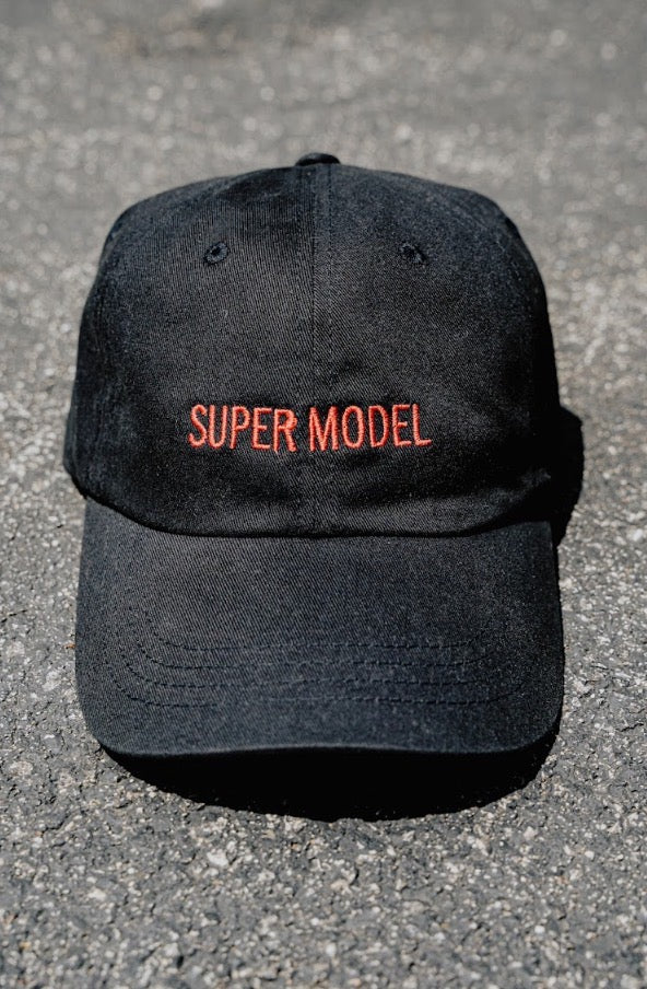 Black cap with 'SUPER MODEL' text on a gray pavement background