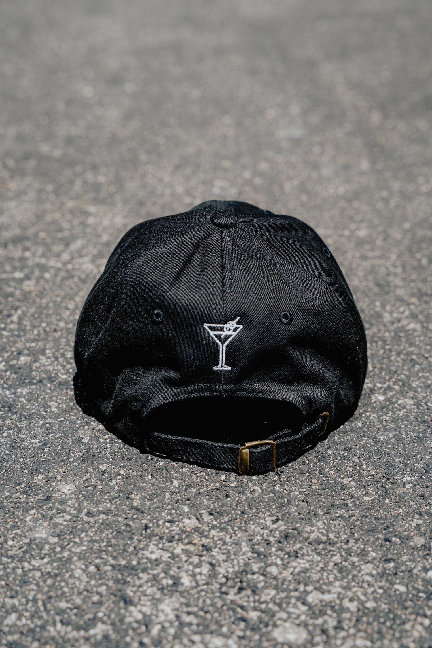 Black cap with a martini glass emblem on a gray pavement background