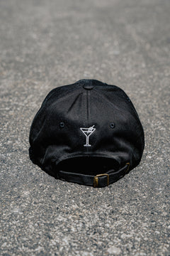 Black cap with a martini glass emblem on a gray pavement background