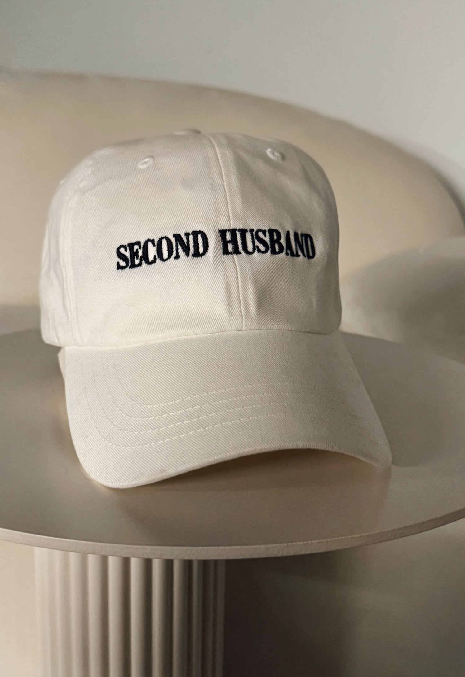 White cap with 'SECOND HUSBAND' text on a beige surface