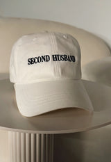 White cap with 'SECOND HUSBAND' text on a beige surface