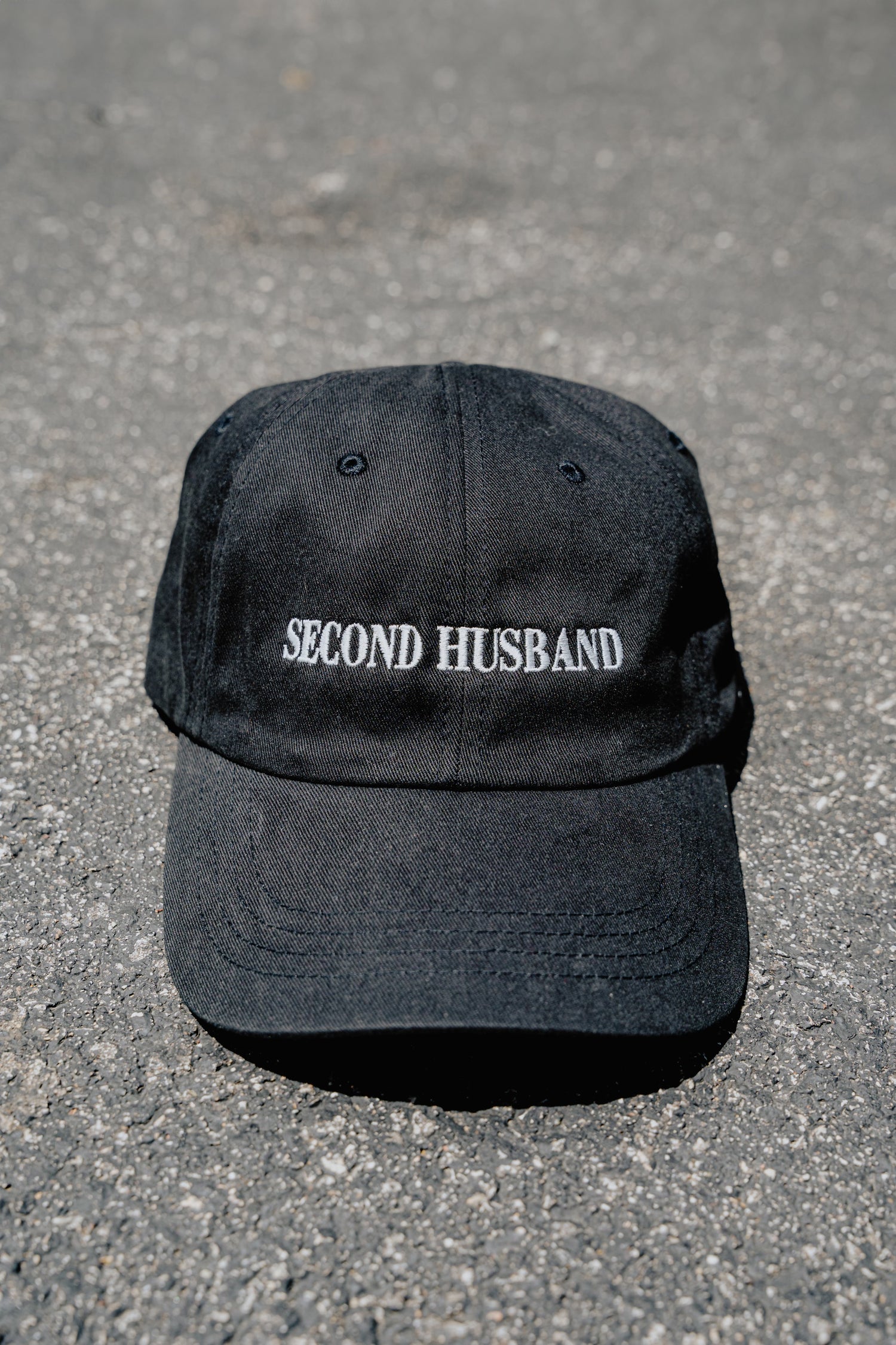 Black cap with 'SECOND HUSBAND' text on a gray concrete surface